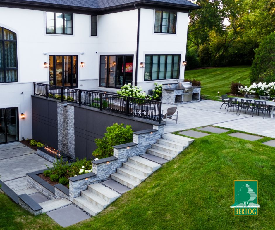 Two-level backyard on a slope with tiered stone retaining walls, lit stone steps, an outdoor kitchen and a lower patio with a linear fire table.