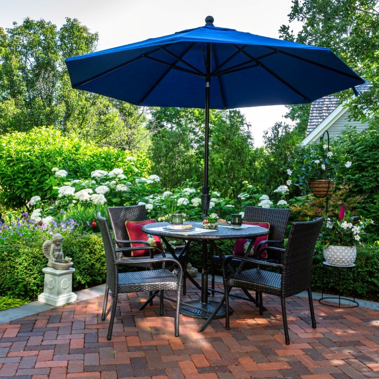 Brick patio with a wicker dining set under a blue umbrella, surrounded by white hydrangeas.