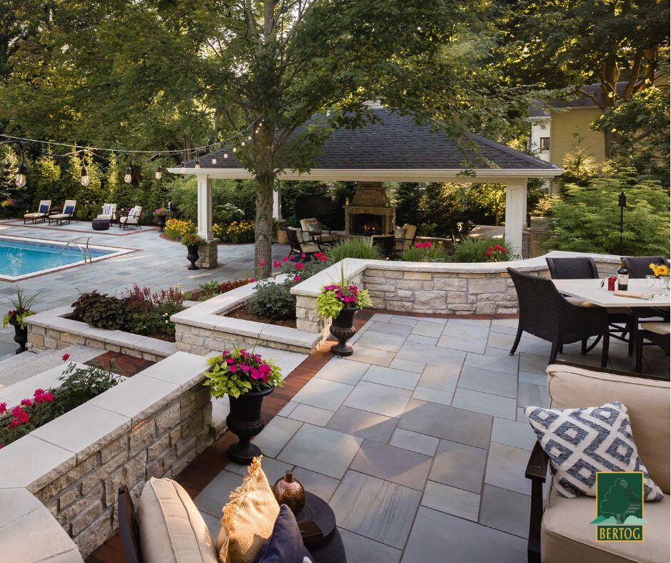 Bluestone patio with stone seat walls, planters of pink flowers, a dining set and a covered pavilion with fireplace beside the pool.