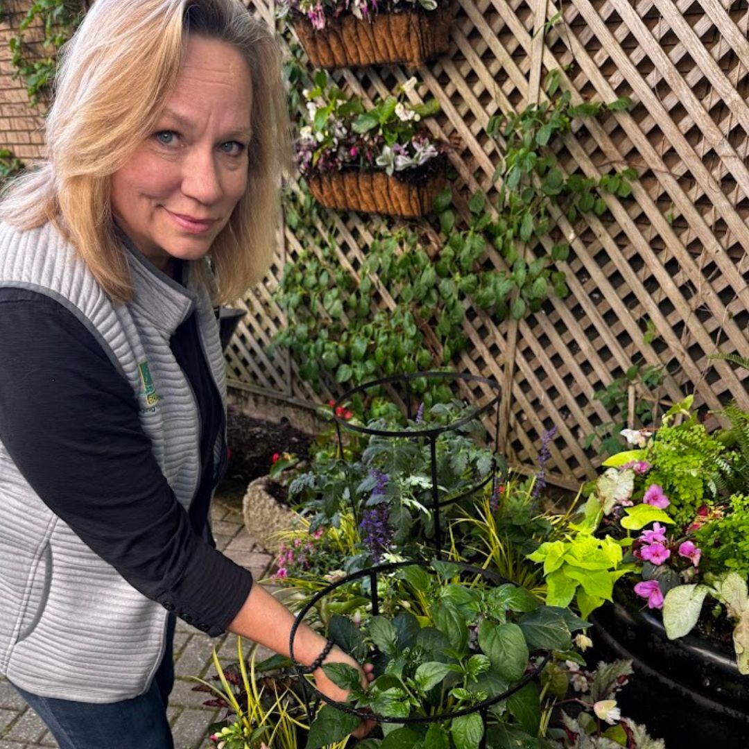 Sue Ryba planting a container of herbs and flowers in front of a lattice wall with hanging baskets.