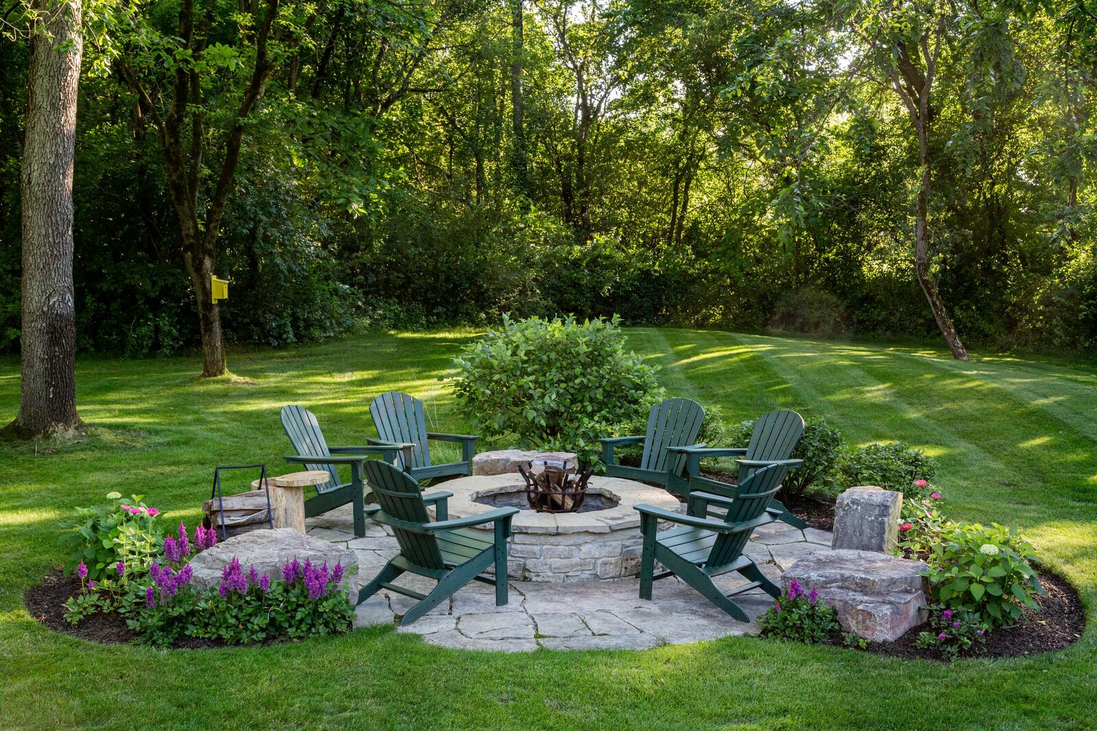 Round stone fire pit on a flagstone patio surrounded by green Adirondack chairs and a freshly striped lawn.
