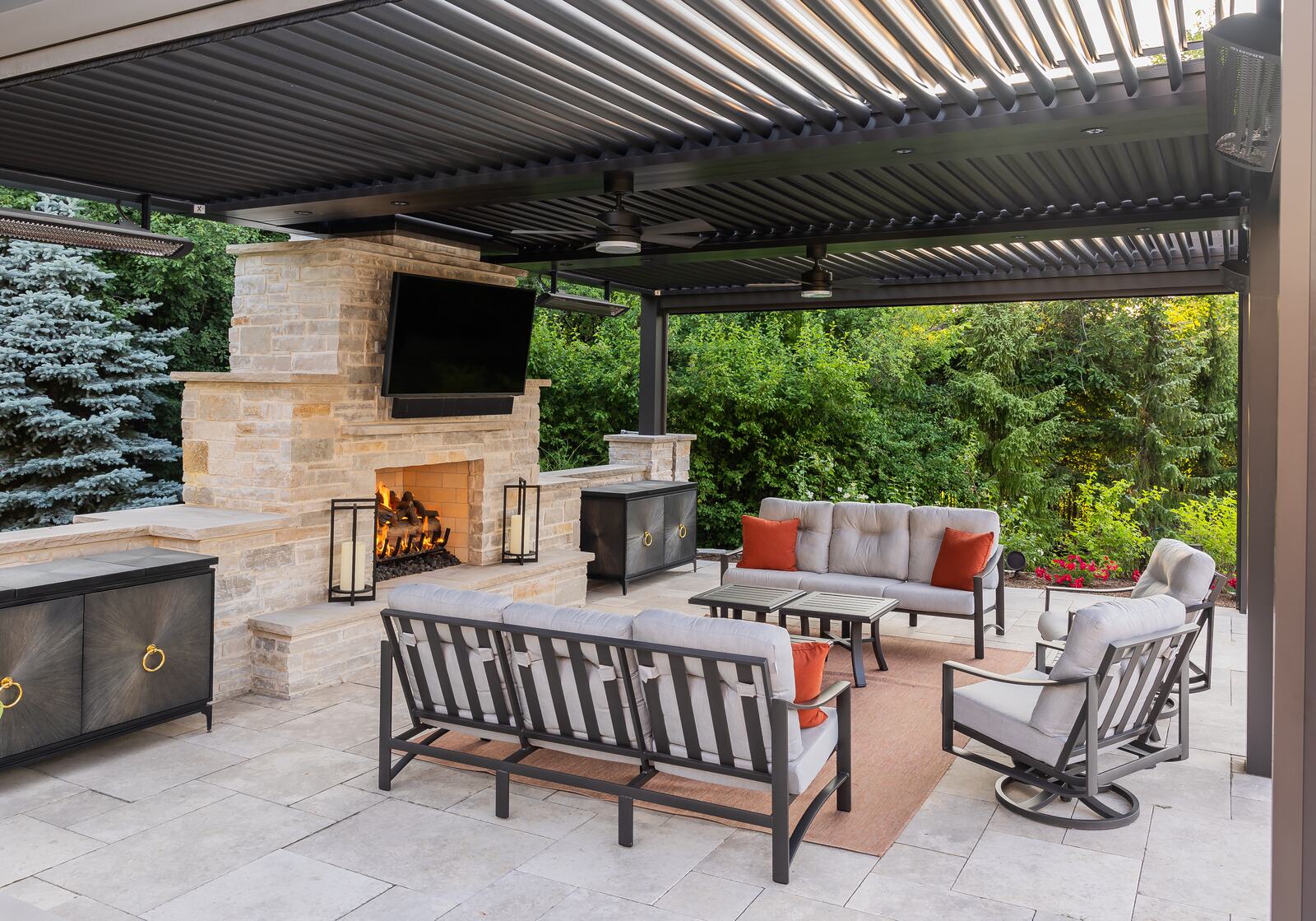 Patio under a louvered pergola with a stone fireplace, mounted TV and gray outdoor sofas with orange pillows.