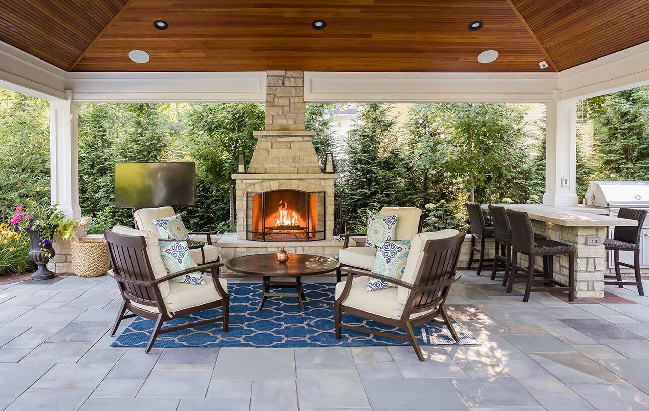 Covered patio pavilion with a stone fireplace, cushioned armchairs and a blue patterned rug.