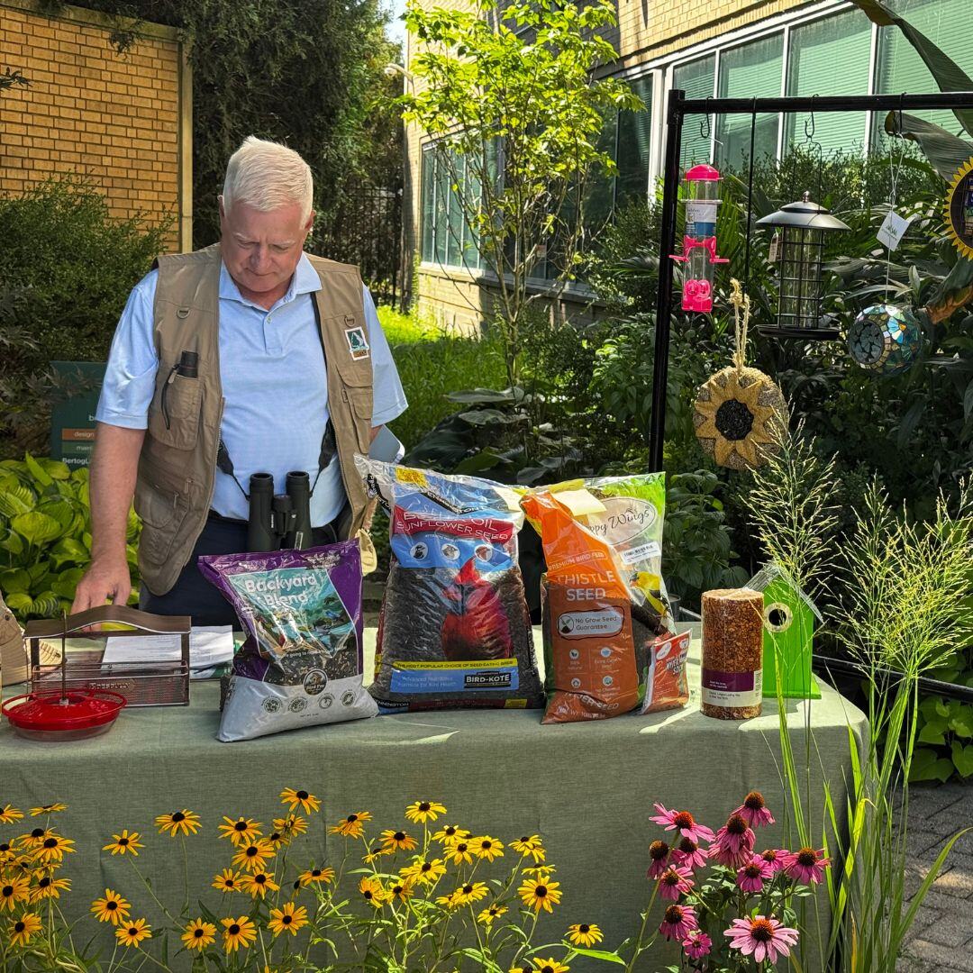 Bob Bertog at an outdoor table of birdseed bags and feeders in front of a garden.