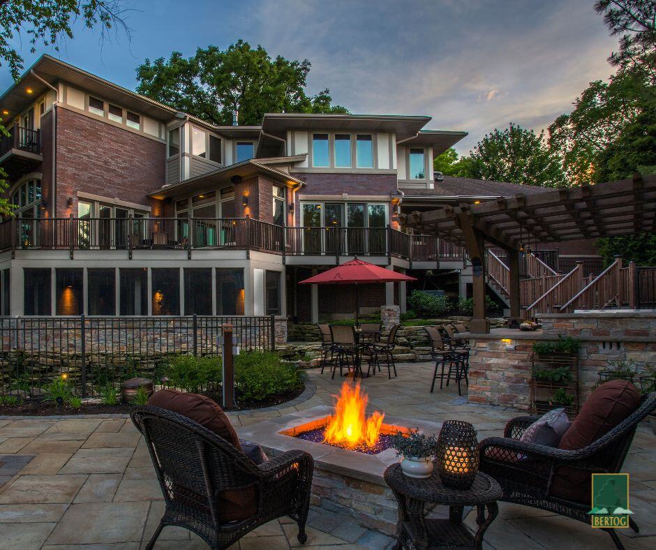 Stone patio at dusk with a lit fire pit, wicker lounge chairs, a dining area under a red umbrella and a pergola beside the house.