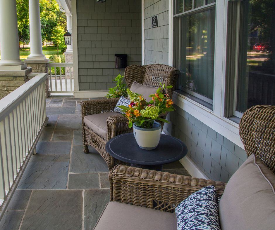 Covered front porch with bluestone floor, wicker armchairs, a round side table with a potted flower arrangement and white railings.